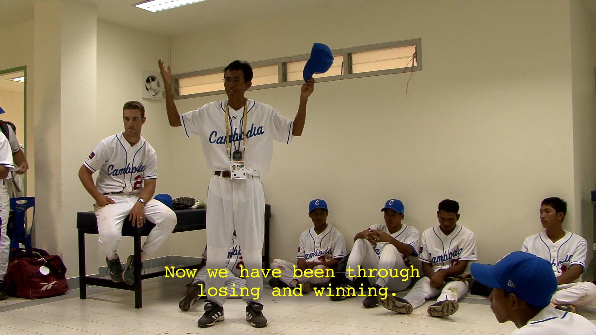 Members of the Cambodia national baseball team reflect on a recent loss against Malaysia before discussing strategies for their upcoming game. The players share a moment of sportsmanship on the field before gathering in the locker room for a team huddle.