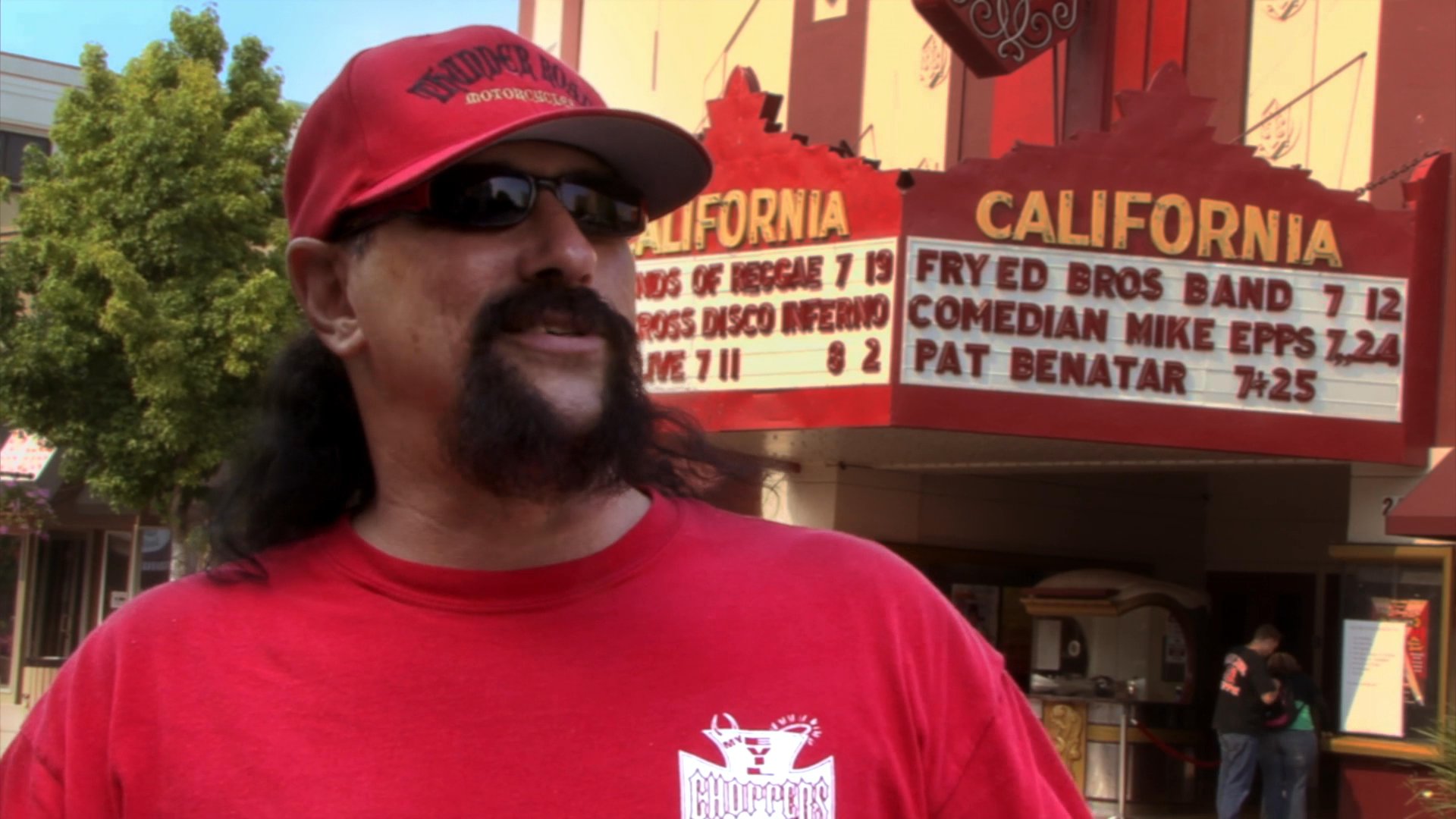 Musician and biker Charlie Brechtel discusses his work during an interview, followed by performance highlights from a stage show in front of a California theater marquee.