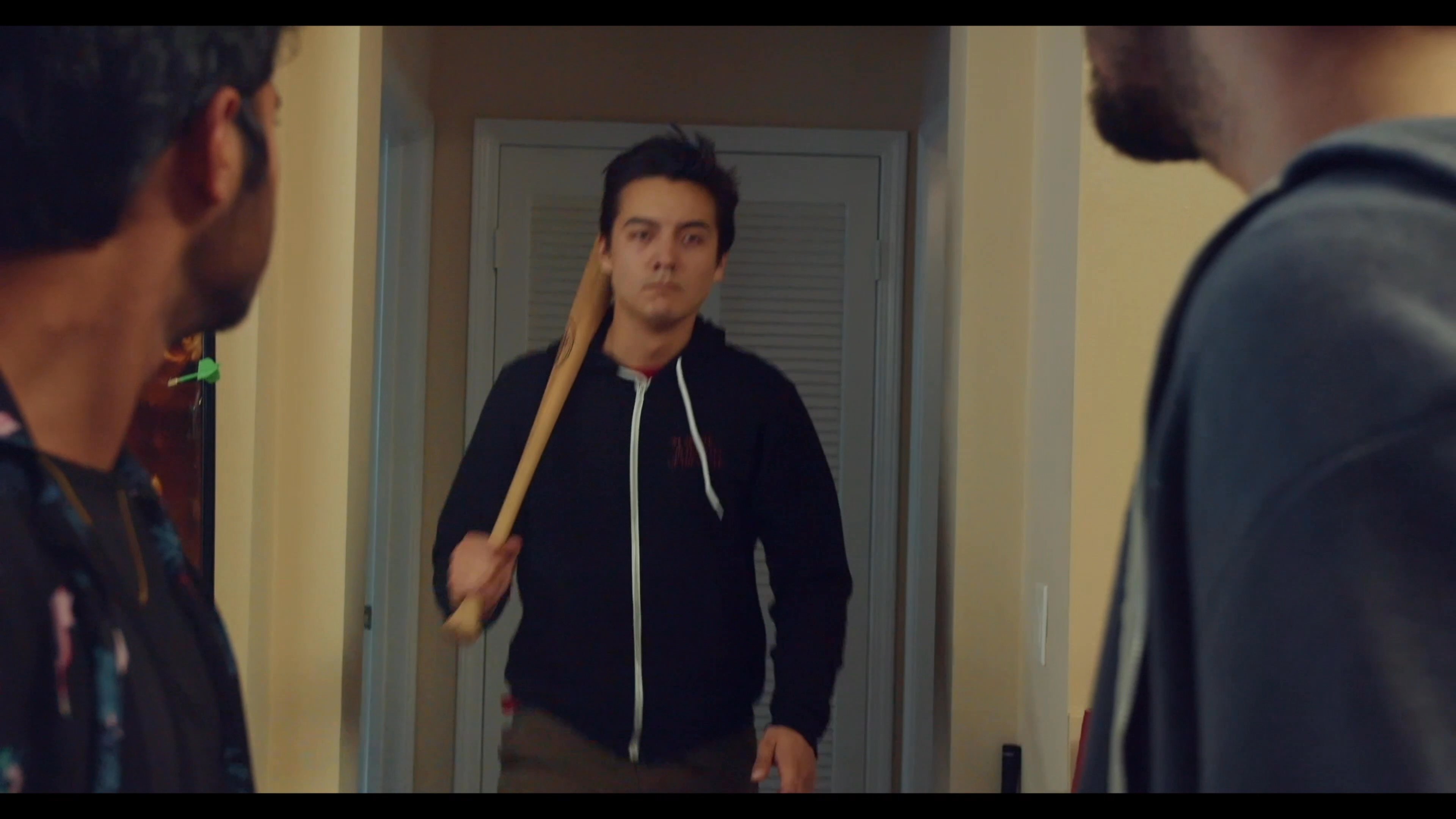A group of young men engage in a tense verbal confrontation inside a residential living room, punctuated by the arrival of an individual carrying a baseball bat.