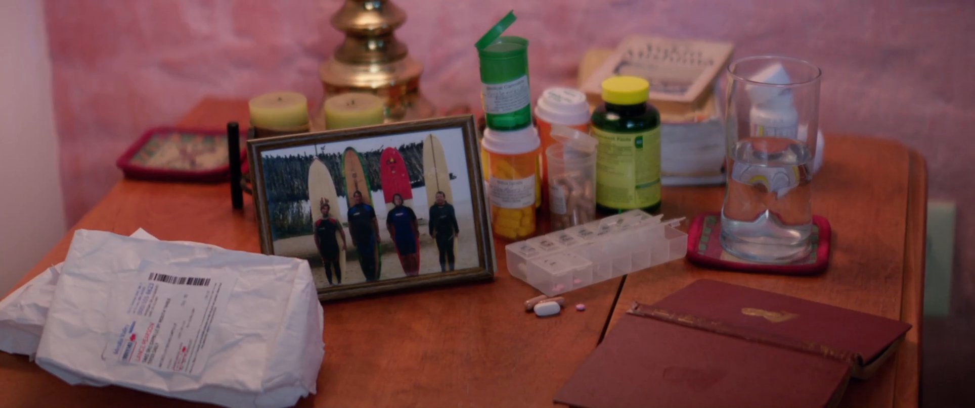 A man observes his surroundings in a quiet room, eventually approaching a table filled with personal belongings and medication. He carefully picks up a small framed photograph, reflecting on the moment in his home environment.