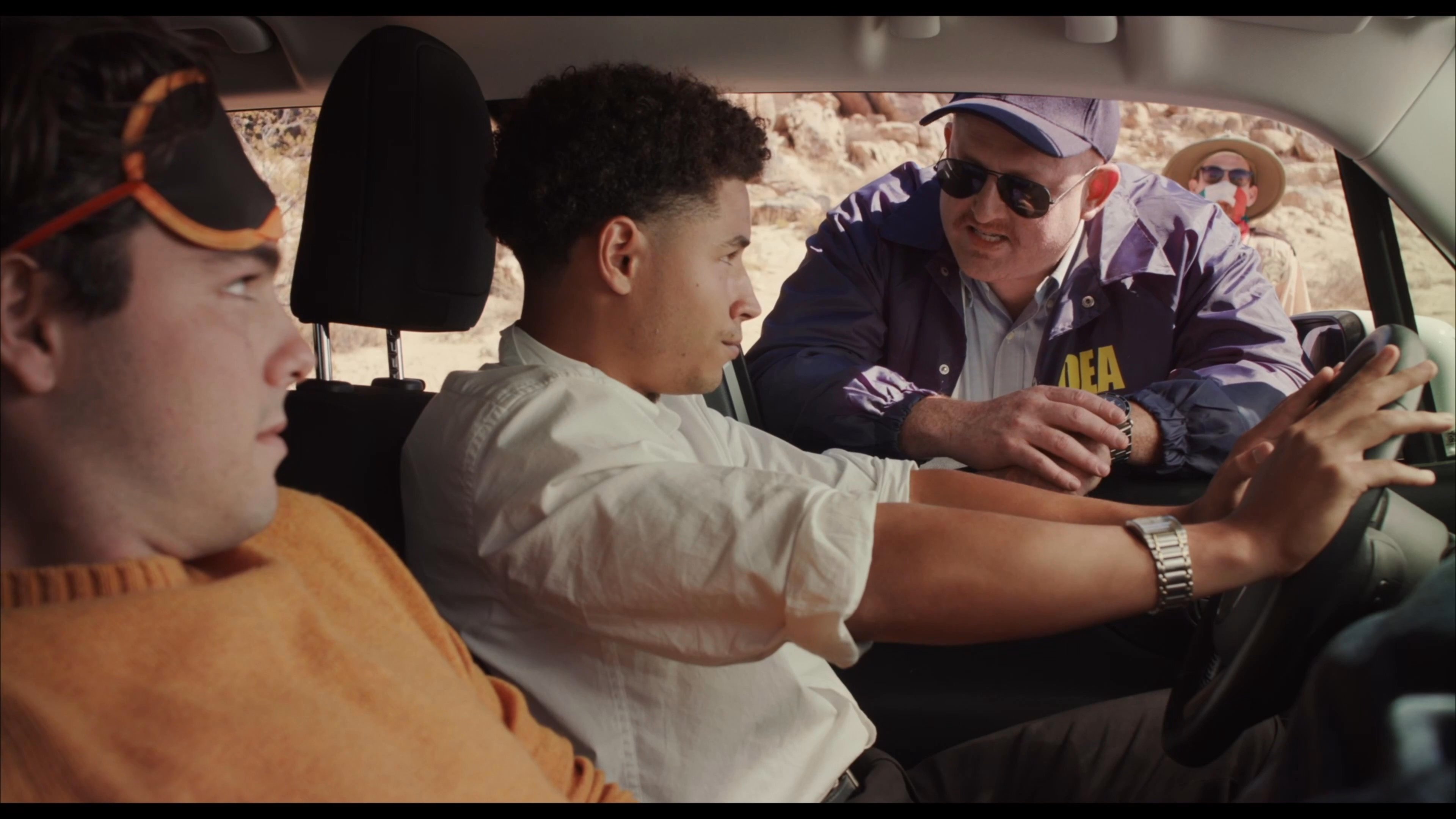 A DEA agent questions two suspects inside a parked vehicle in a remote desert environment. The agent maintains an authoritative tone while observing the individuals.