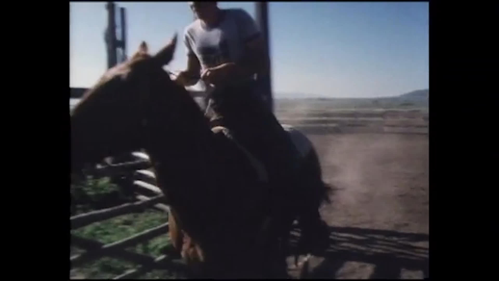 A rider circles a horse around a dusty fenced arena while an older man recalls carrying young Jeff on the saddle. A dog crosses the yard as the man stands beside the horse and gestures toward its saddle.