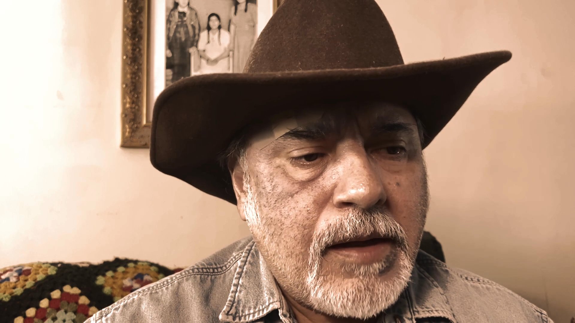 An older man wearing a brown cowboy hat delivers a direct, conversational monologue while seated indoors.