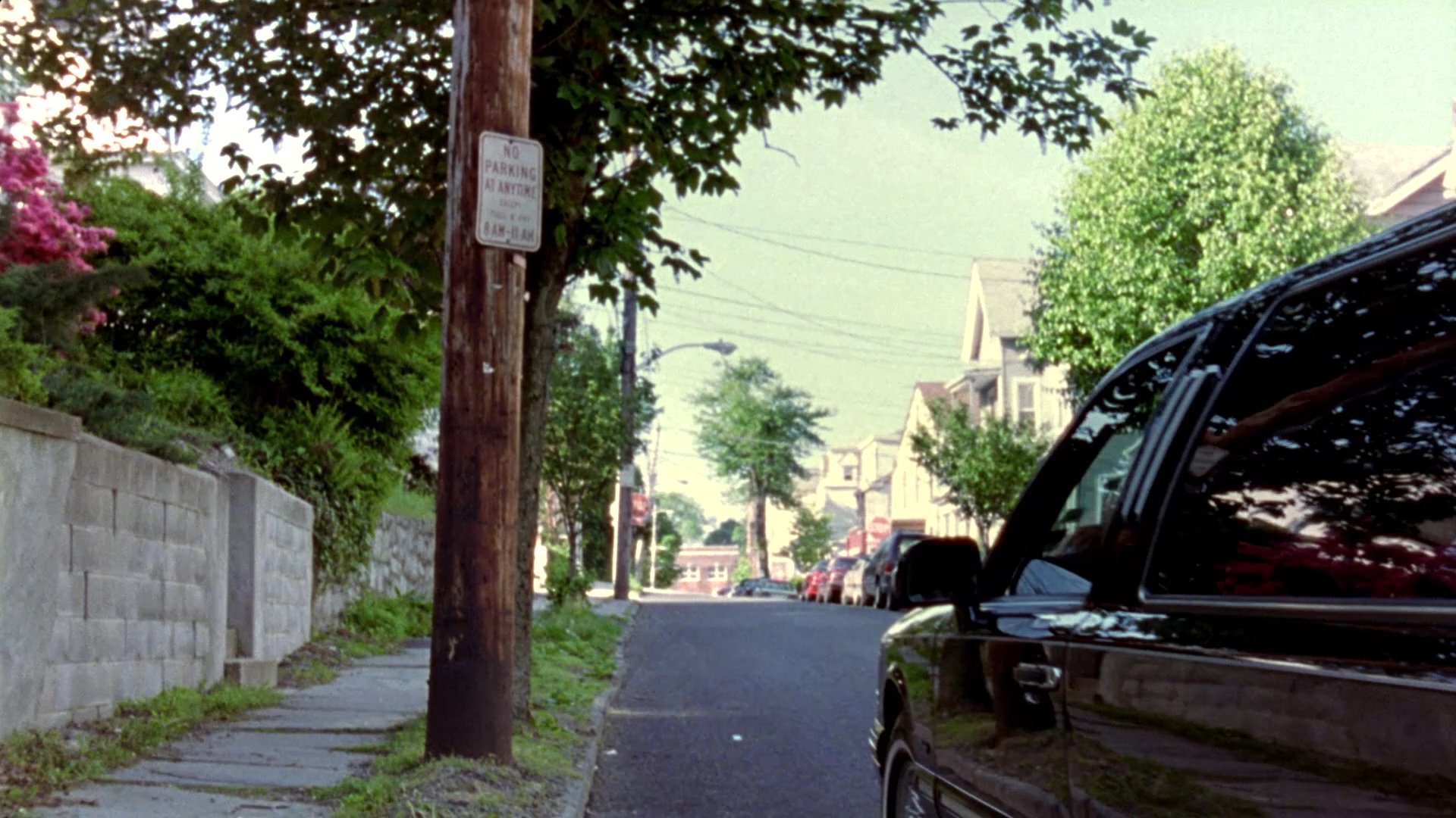 An aerial miniature model of a neighborhood transitions into a ground-level perspective as a black limousine drives down a residential street and stops in front of a house.
