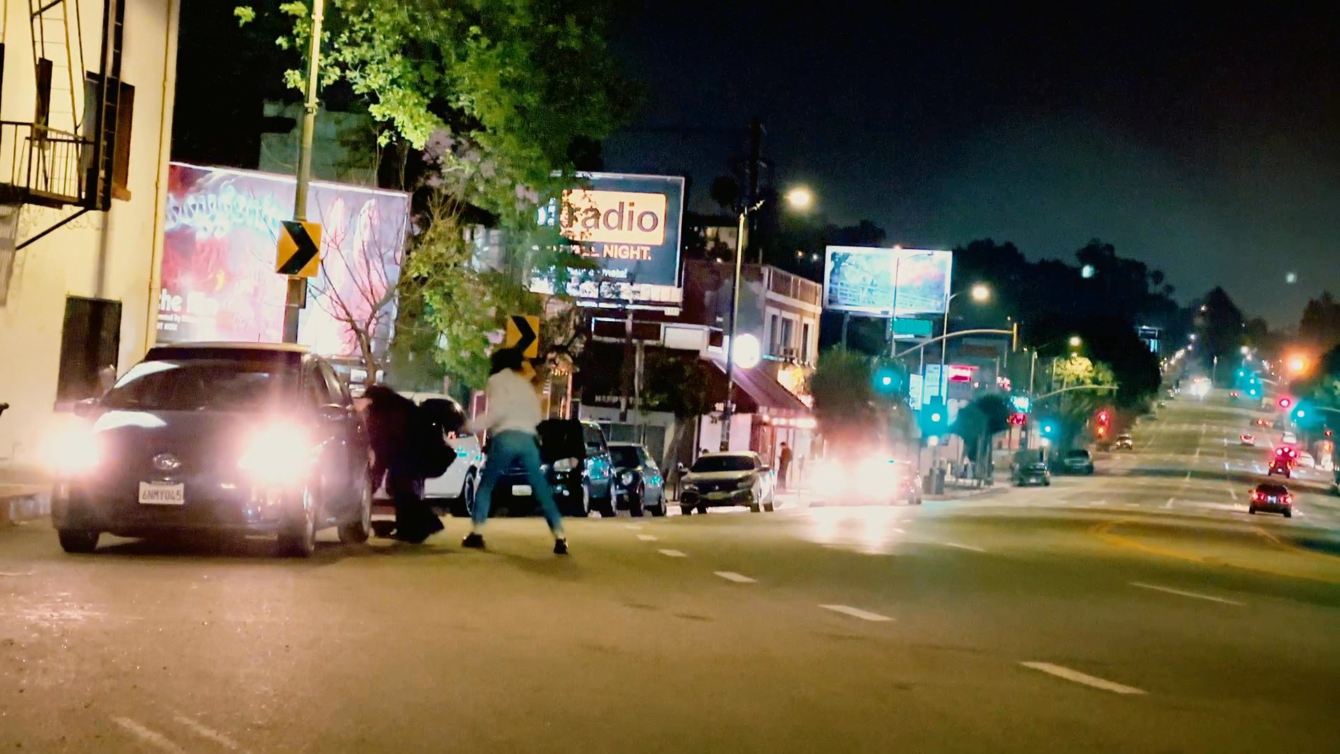 A man exits his car on a dark city street, initiating an interaction with pedestrians nearby while two passengers watch from the vehicle. The scene captures a tense nocturnal urban encounter.