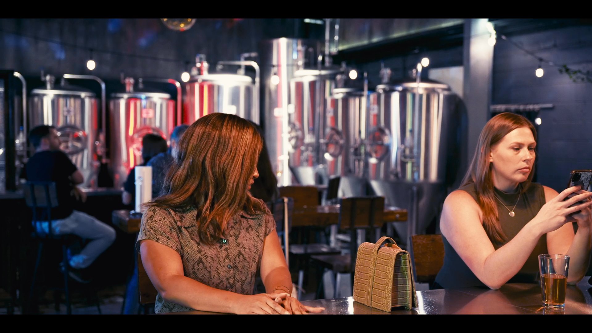 A bartender interacts with patrons at a brewery counter, serving drinks in an industrial atmosphere. Guests occupy the bar area while engaged in casual conversation and social activity.