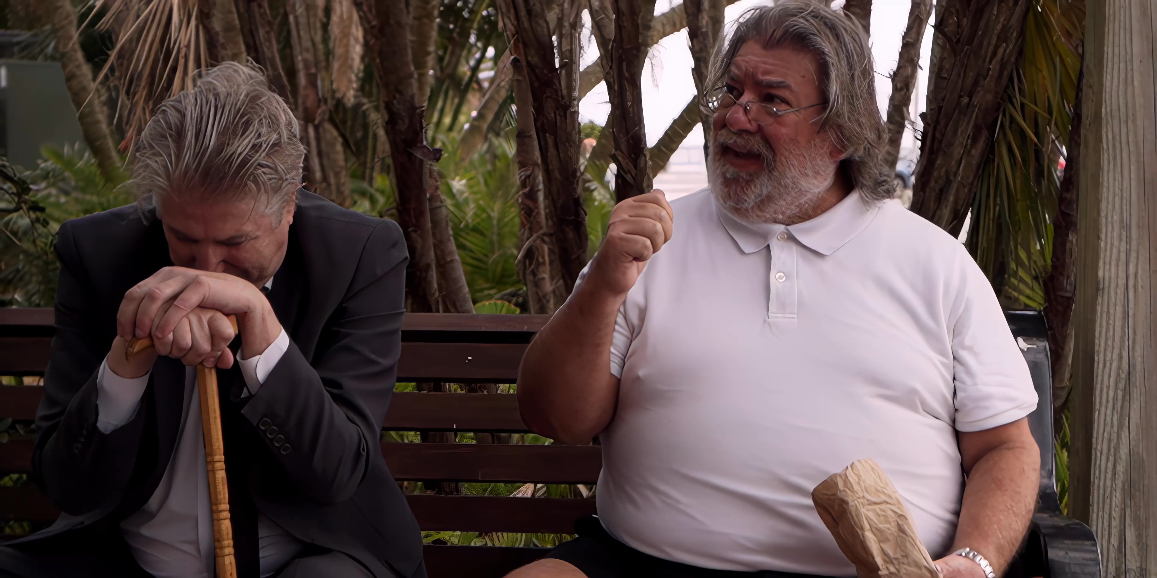 Two middle-aged men engage in a calm conversation while sitting on a park bench surrounded by trees.