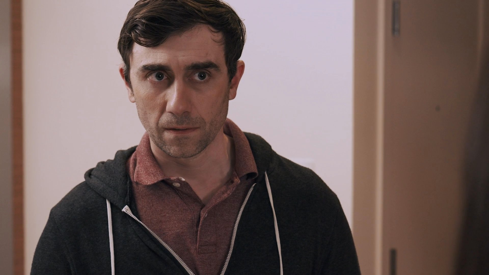 A man and woman engage in a serious, tense conversation within a residential living space. They exchange expressions of concern and frustration while standing in their home.