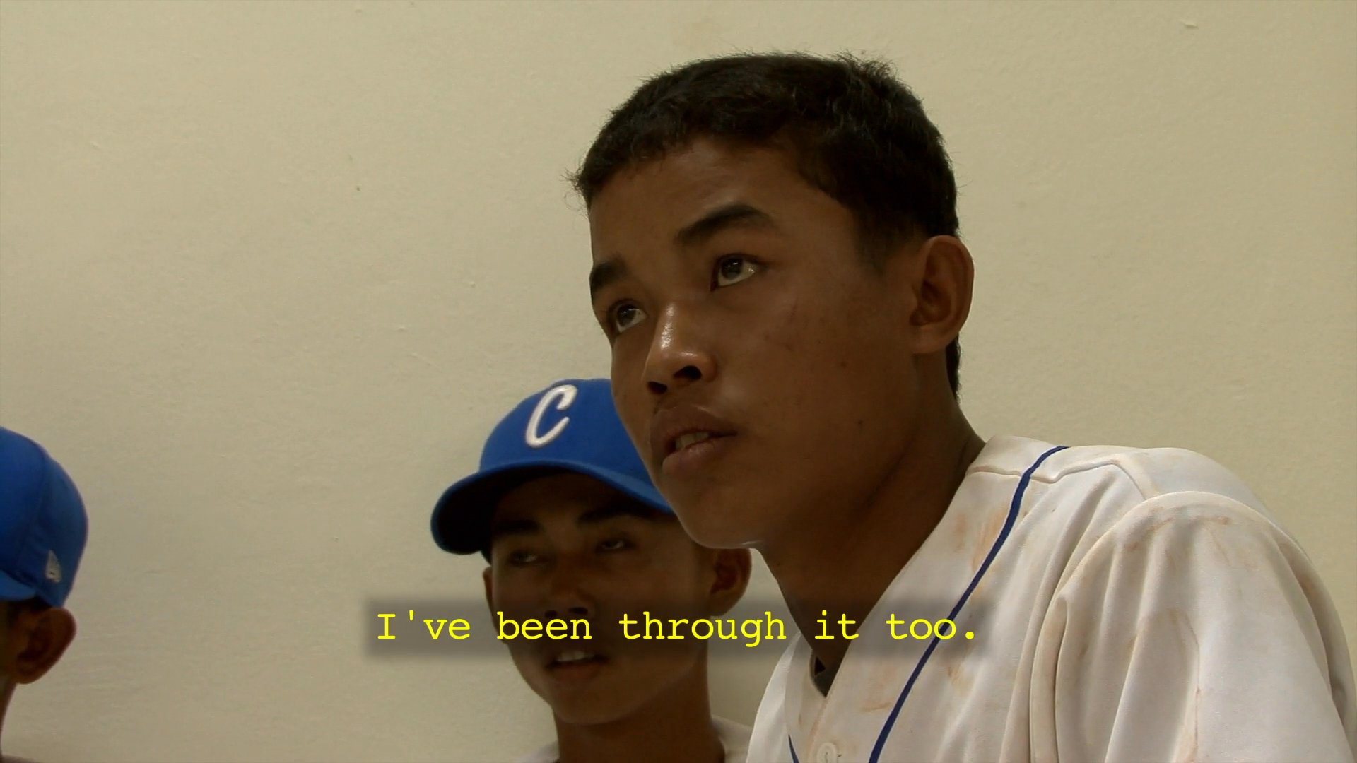 A coach delivers a motivational speech to a Cambodian baseball team in a locker room setting, reflecting on their shared experiences and national pride.