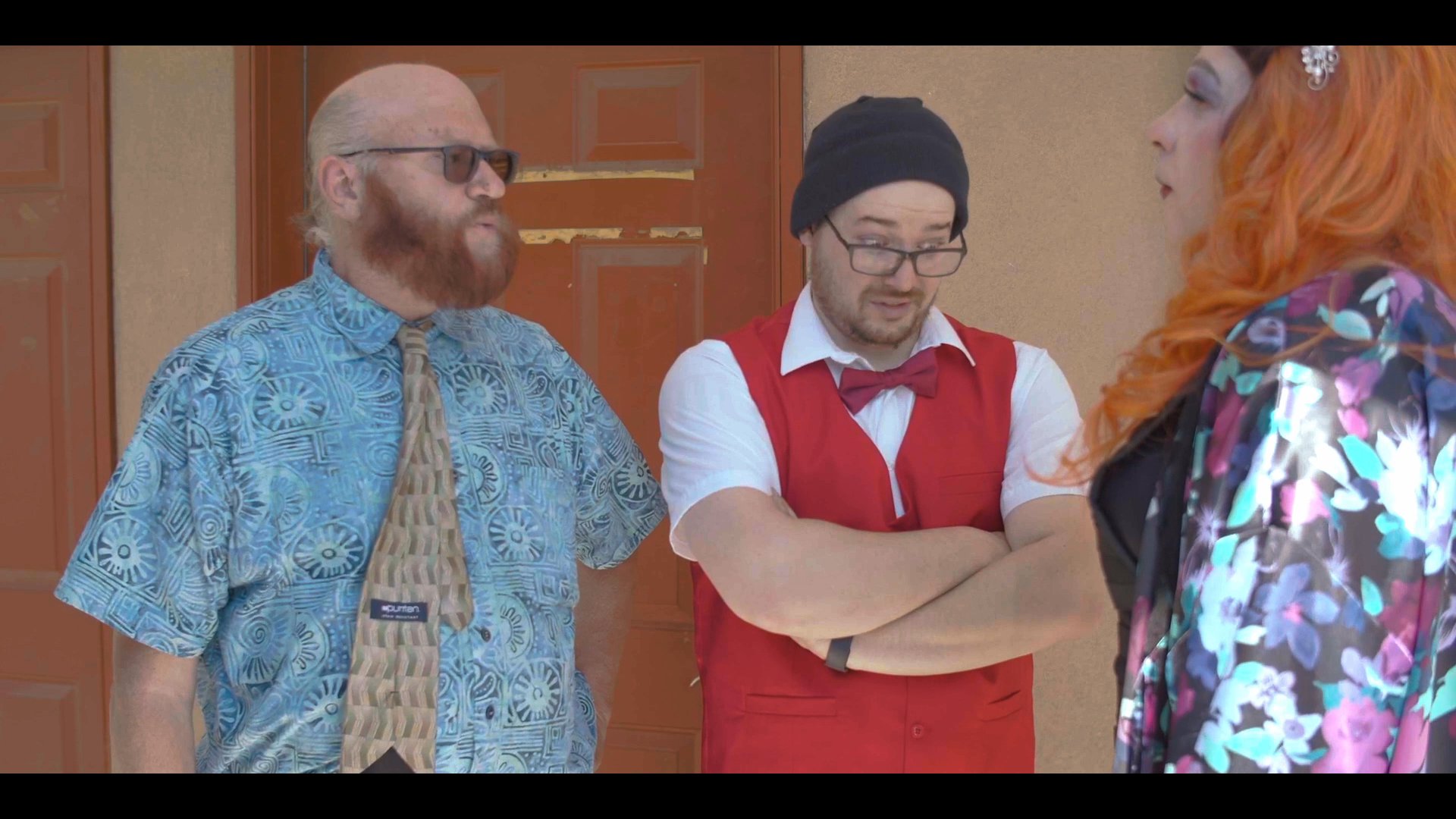 A drag performer in a vibrant floral robe and orange wig exits a vehicle at a motel and initiates a conversation with two men.