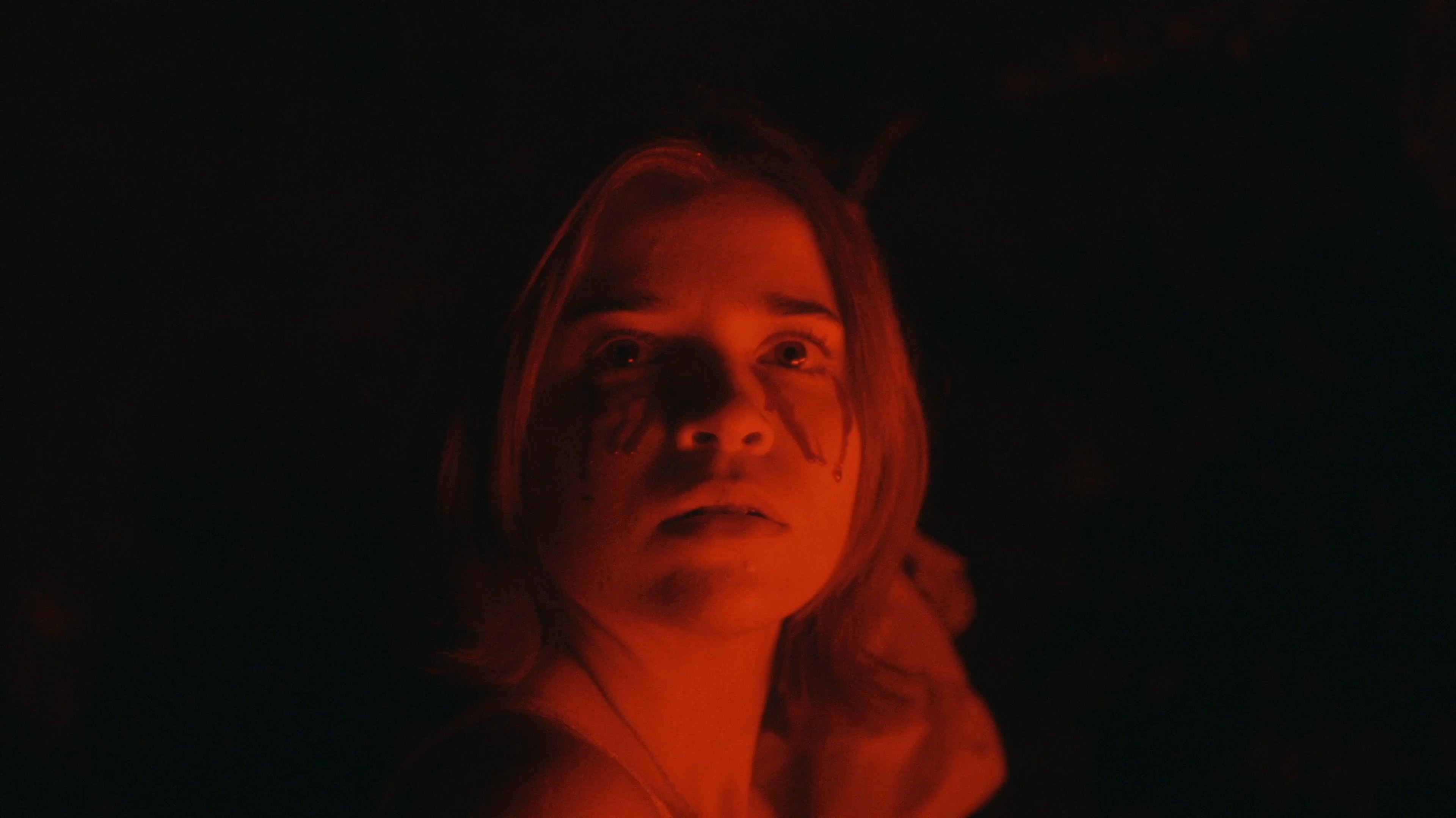 A woman with tear-stained cheeks appears in a dark, red-lit environment, reflecting intense emotional distress. She slowly turns her head, conveying a sense of vulnerability and isolation.