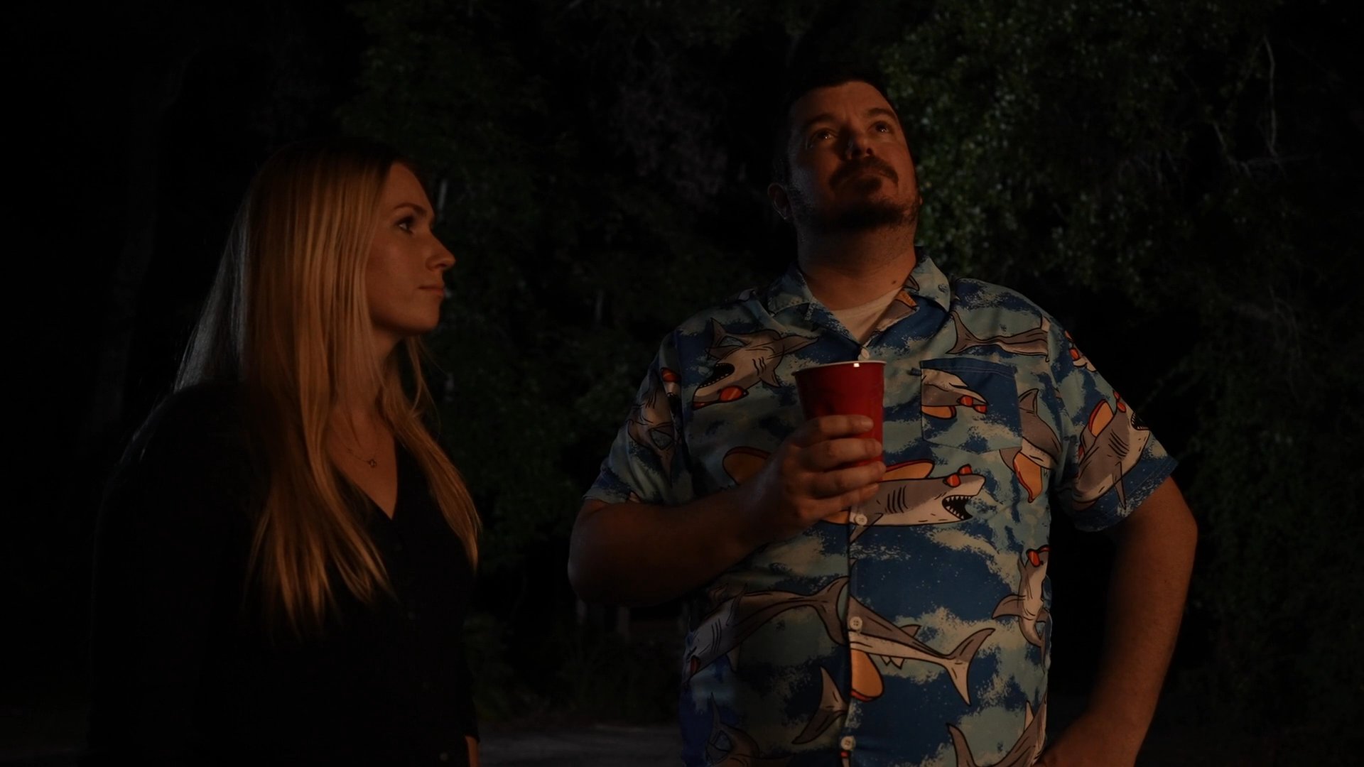 A man and a woman stand outdoors at night, engaging in a quiet conversation. The man holds a drink while both individuals look toward the sky.