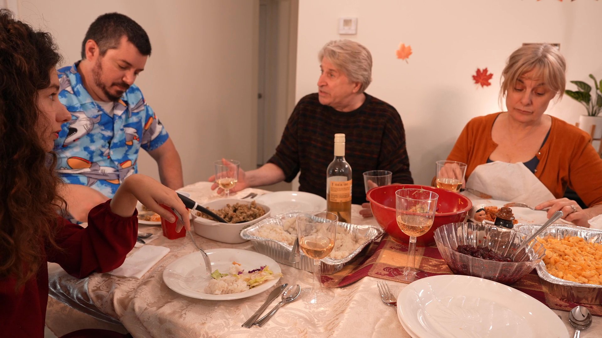 A family prepares a meal in the kitchen and gathers for a communal dinner, sharing conversation around a table.
