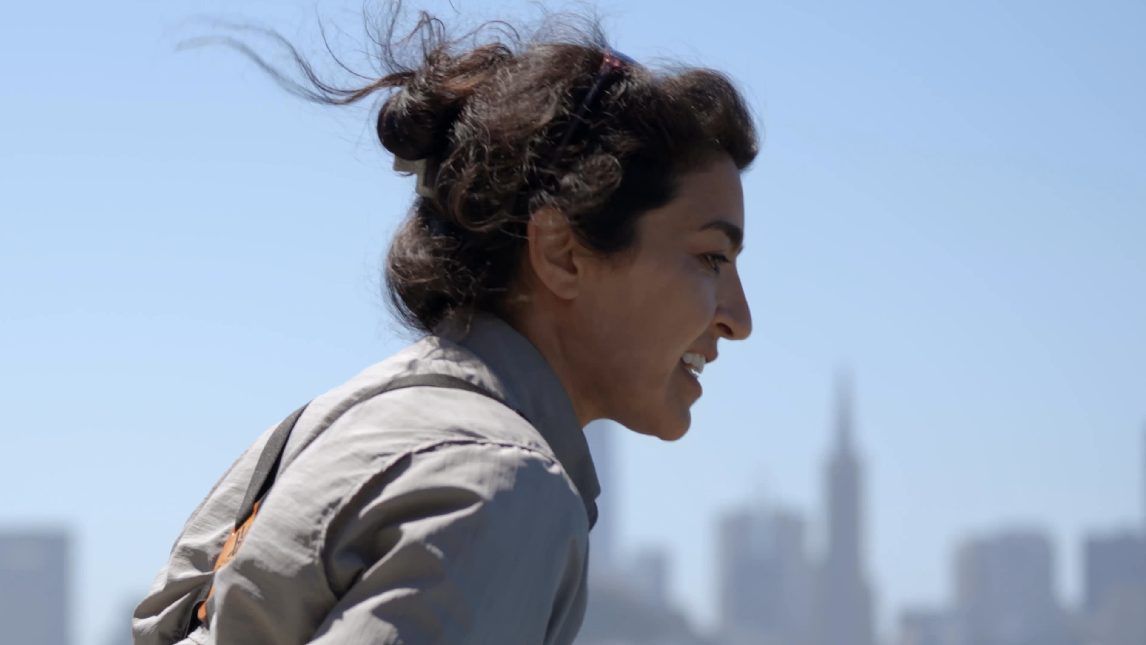 A woman in a gray field uniform discusses her career and the support of her parents, who are originally from Argentina, as the first in her family to attend college. Dark waterbirds swim and flap on the bay, with coastal plants and a distant city skyline in the background.