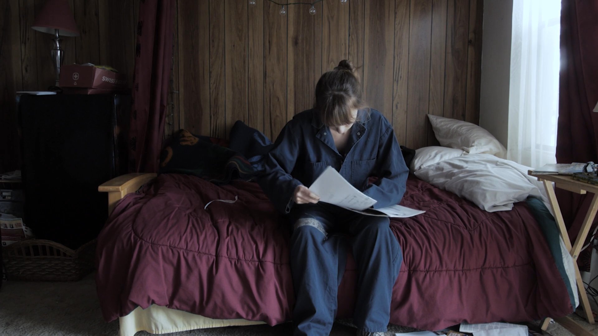 A woman sits on her bed and carefully reads a letter from a bank regarding the settlement of an estate. She appears contemplative and somber while processing the financial information.