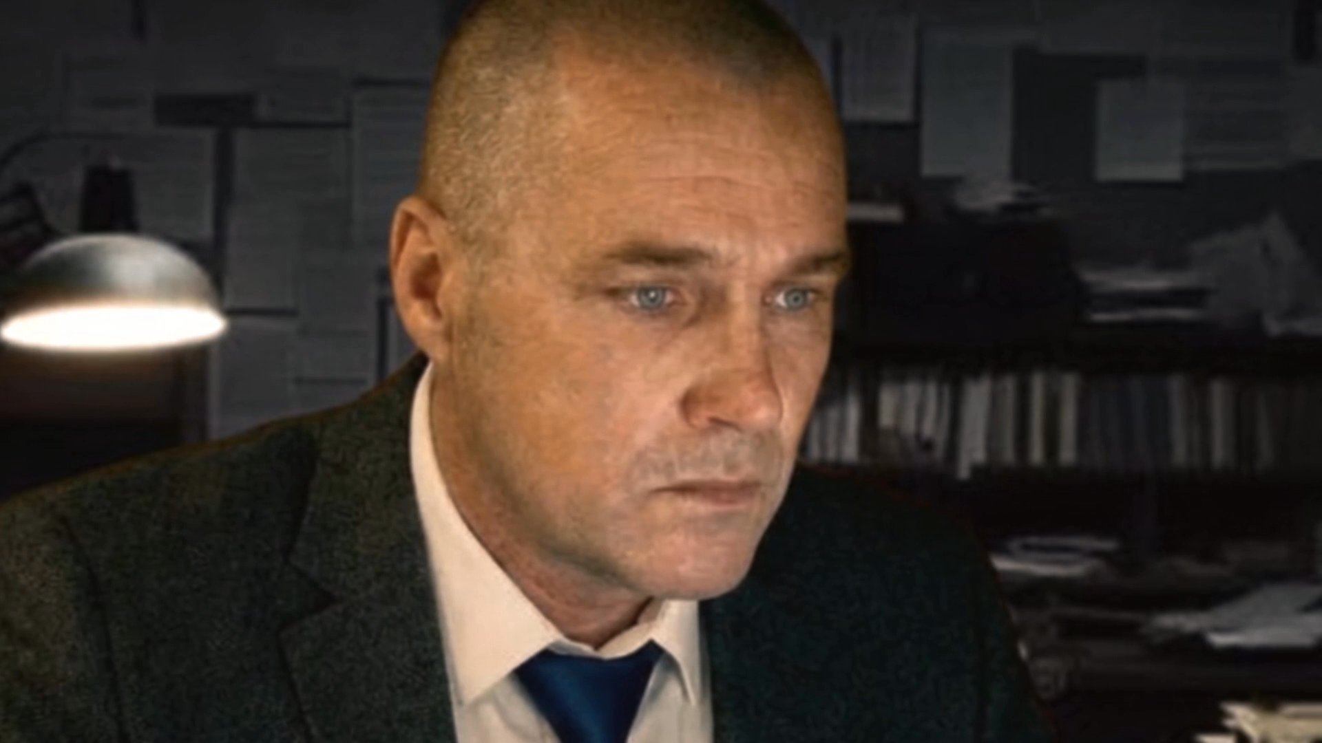 A man in a suit works diligently at his desk in a dimly lit office, surrounded by stacks of case files and a framed family portrait.