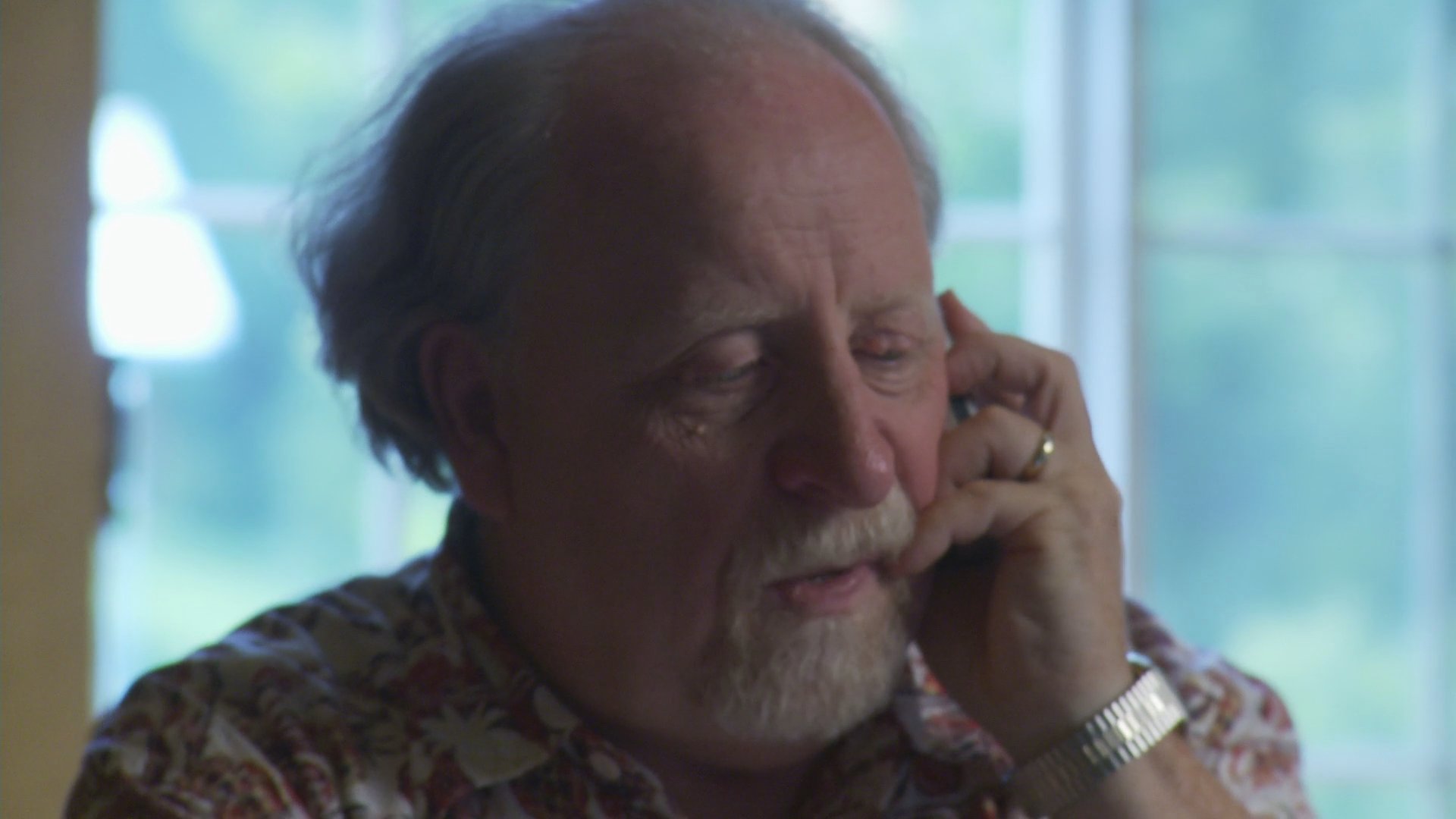 An older man experiences visible distress while speaking on the telephone in a brightly lit room. His facial expressions and gestures indicate he is processing difficult or somber news.