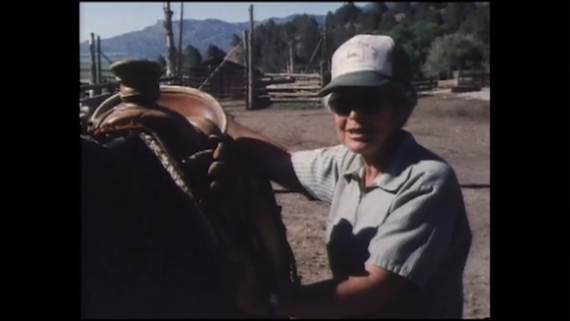 Beside a saddled horse in a ranch corral, a rancher gestures toward the saddle while recalling carrying a child on rides to gather cattle. The story includes a pillow in the seat, a bottle, and how much the child enjoyed going faster.