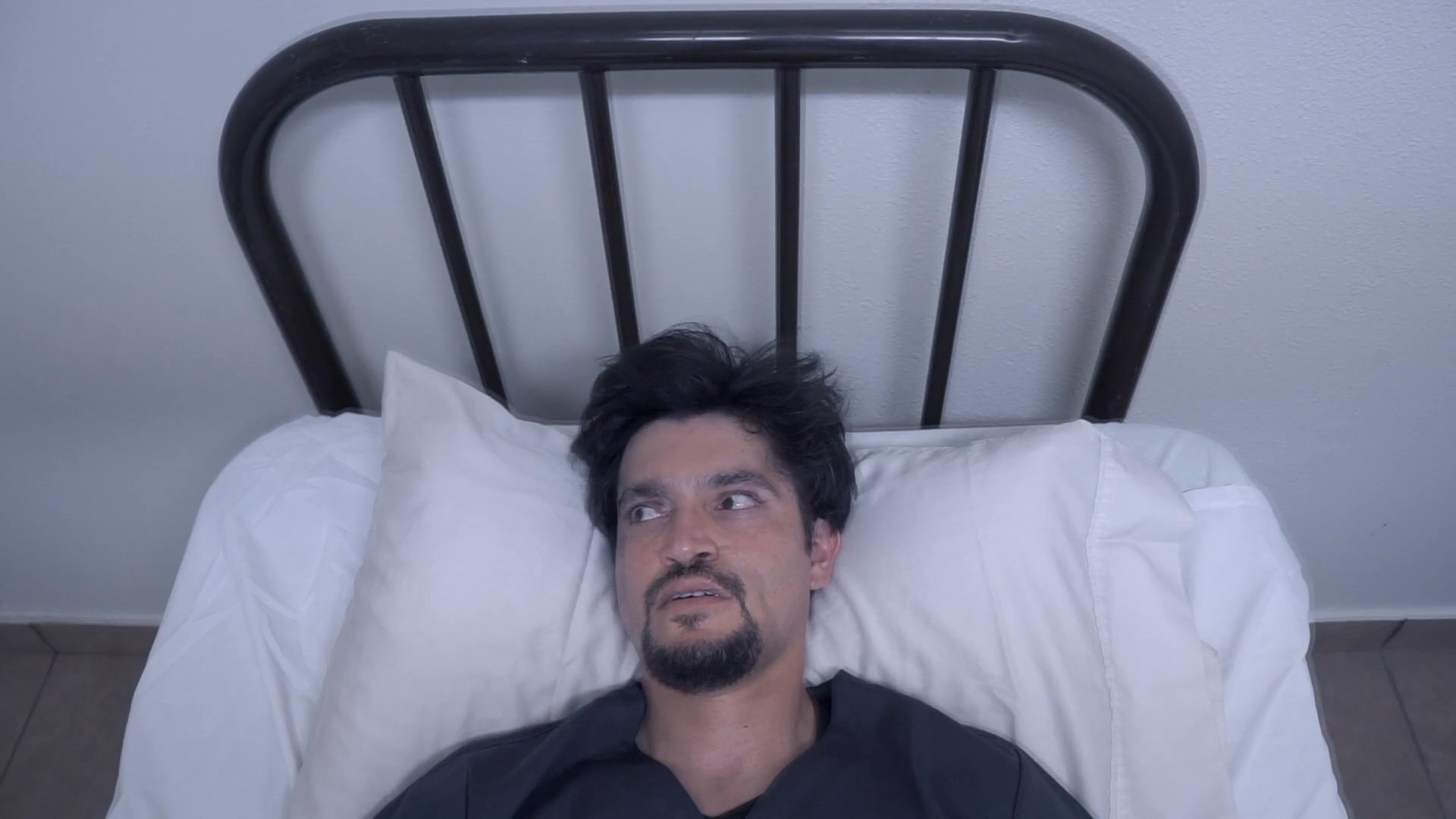 A man lies confined in a stark, sterile hospital room while under the watchful gaze of an observer. The narrative emphasizes themes of psychological distress and the feeling of entrapment.