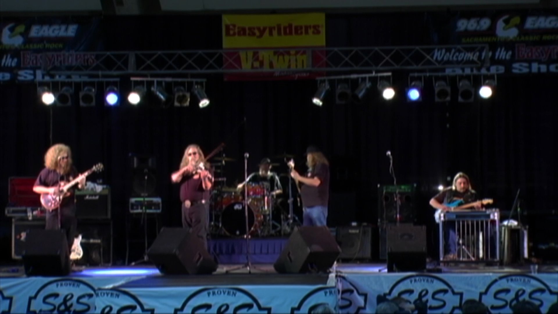 A musician performs a solo on an electric violin while accompanied by a guitarist on a concert stage. The performance captures the interplay between the two instrumentalists in a live event setting.