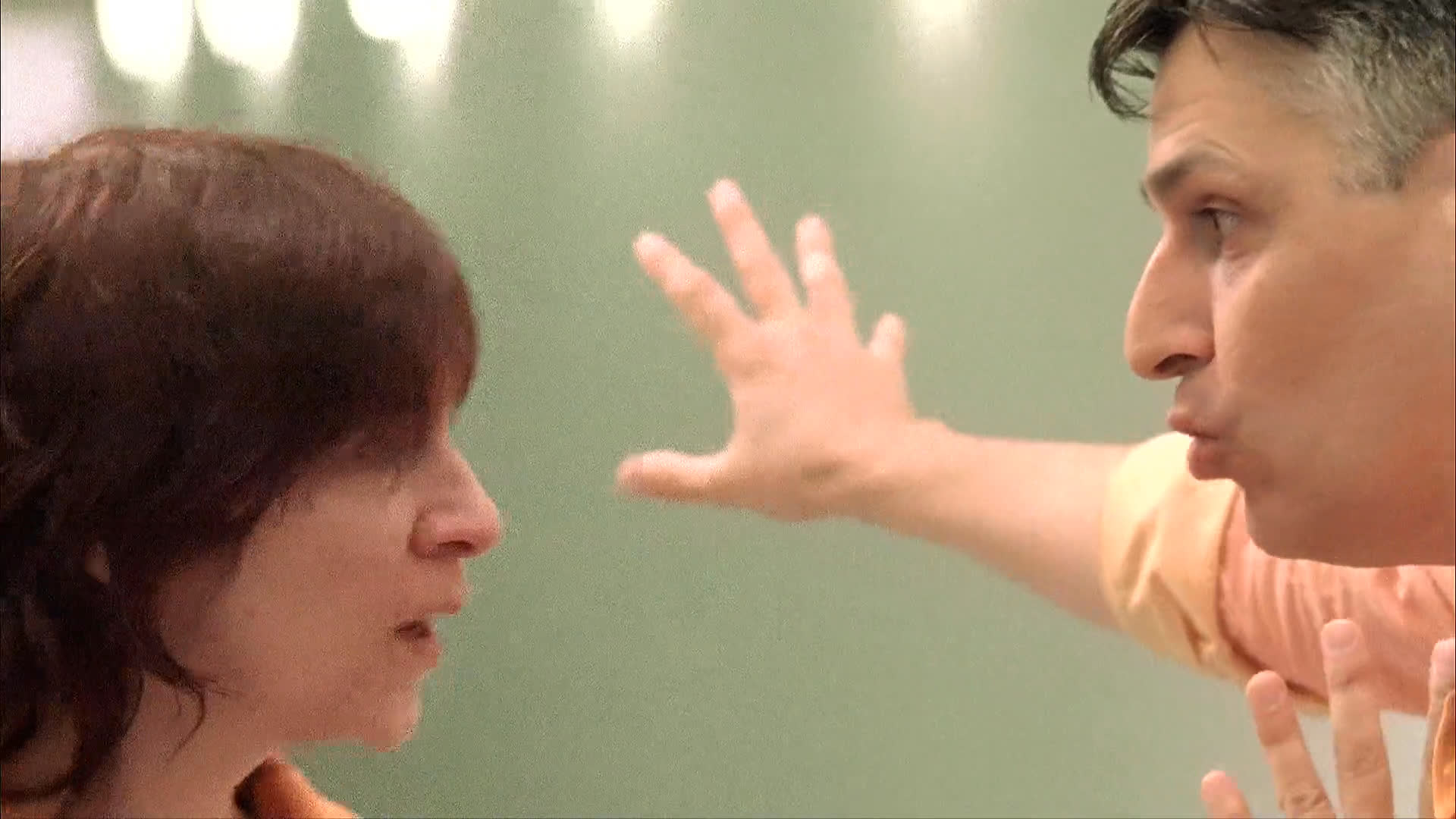 Two colleagues engage in a tense verbal discussion within a brightly lit office environment, characterized by expressive gestures and direct communication.