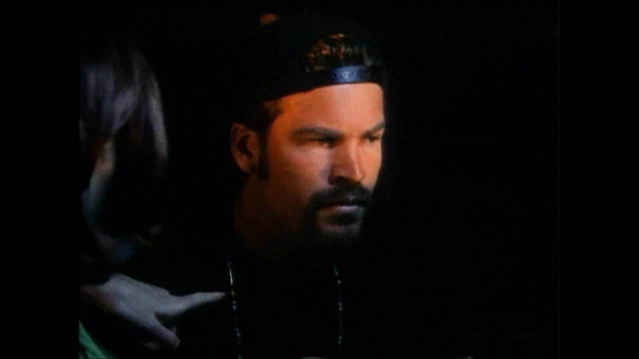 A man and a woman engage in a serious, low-light conversation, focusing on facial expressions and interpersonal tension.