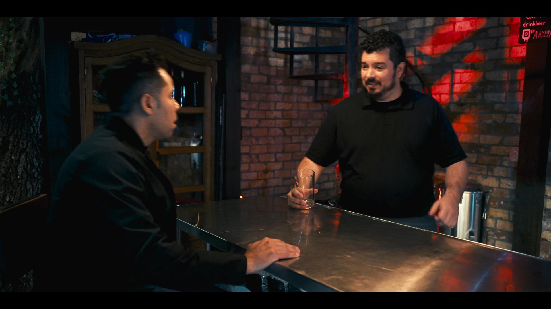 Two men enter a dimly lit, rustic bar setting to engage in a formal, private conversation across a metal counter. The exchange involves focused dialogue and expressive gestures as they discuss their subject matter.