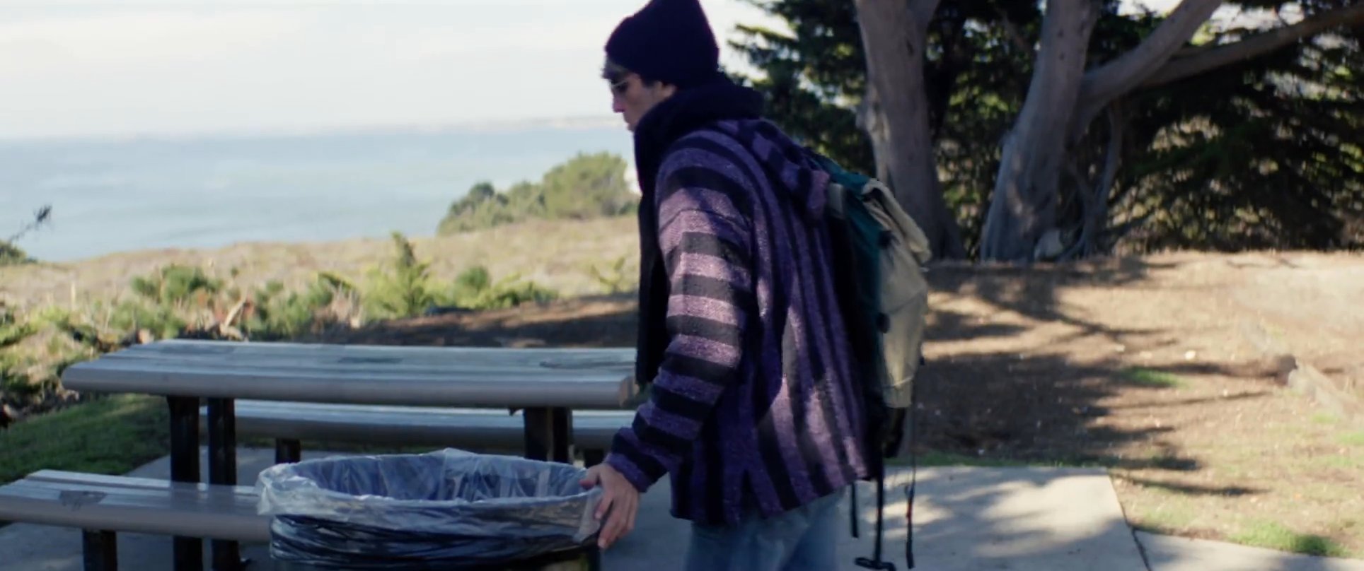 Two surfers observe ocean conditions from a park area before one of them disposes of litter in a trash bin.