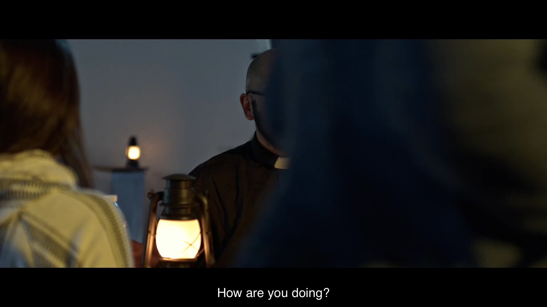 Father Andres introduces himself to Carlos and Maricela during a quiet gathering in a dimly lit community hall. The trio engages in a polite, formal exchange of greetings.