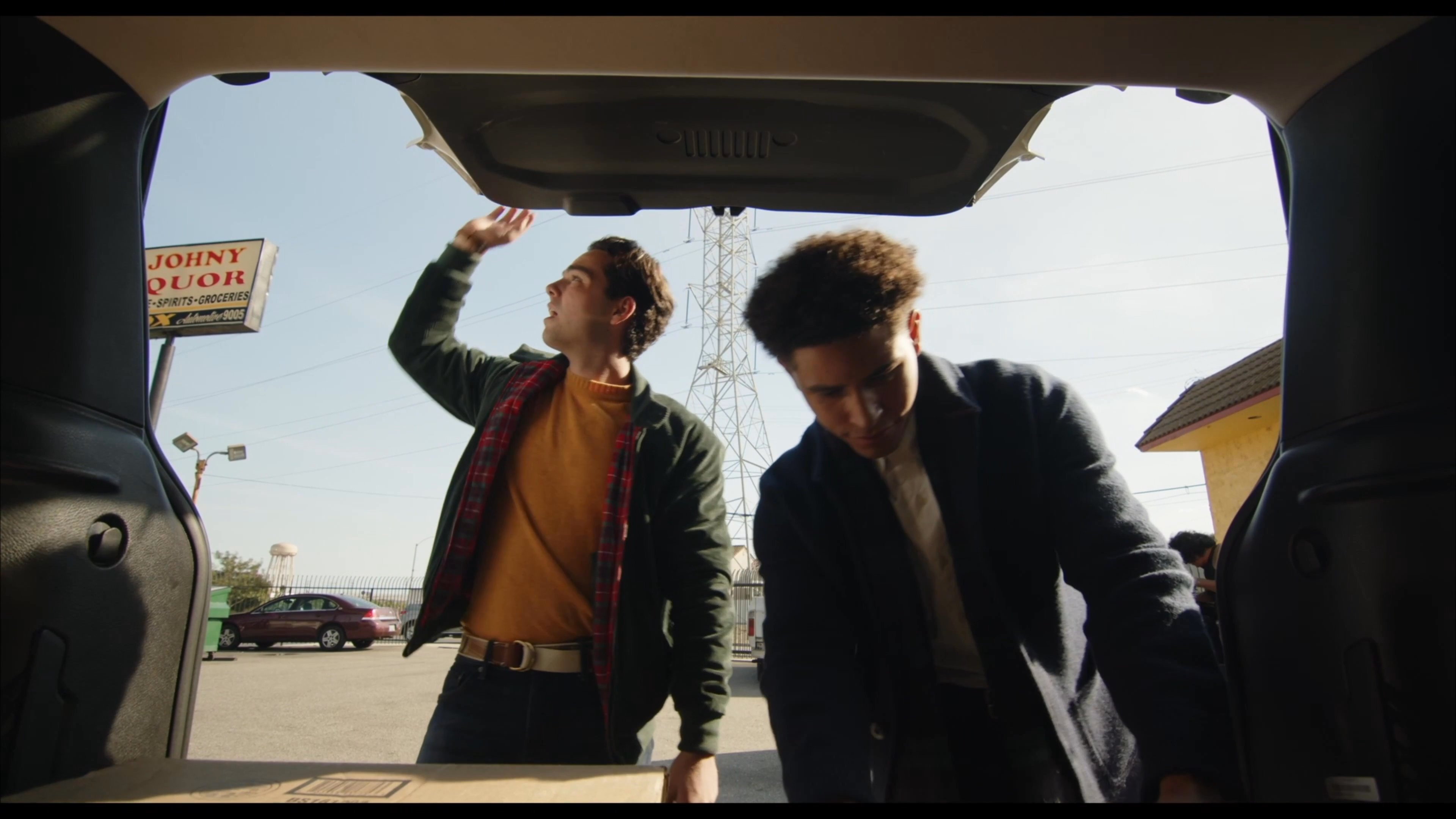 Two men exit a media rental store and load boxes into the trunk of a vehicle in an outdoor parking lot.