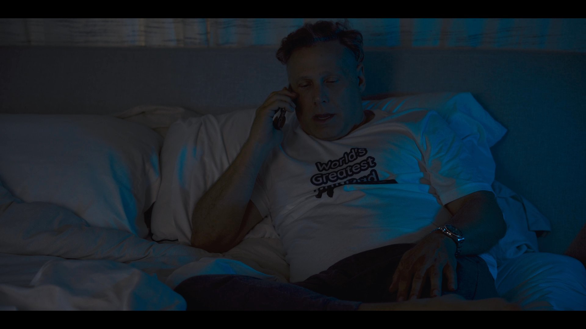 A man wearing a t-shirt reclines in bed at night, engaged in a phone conversation.