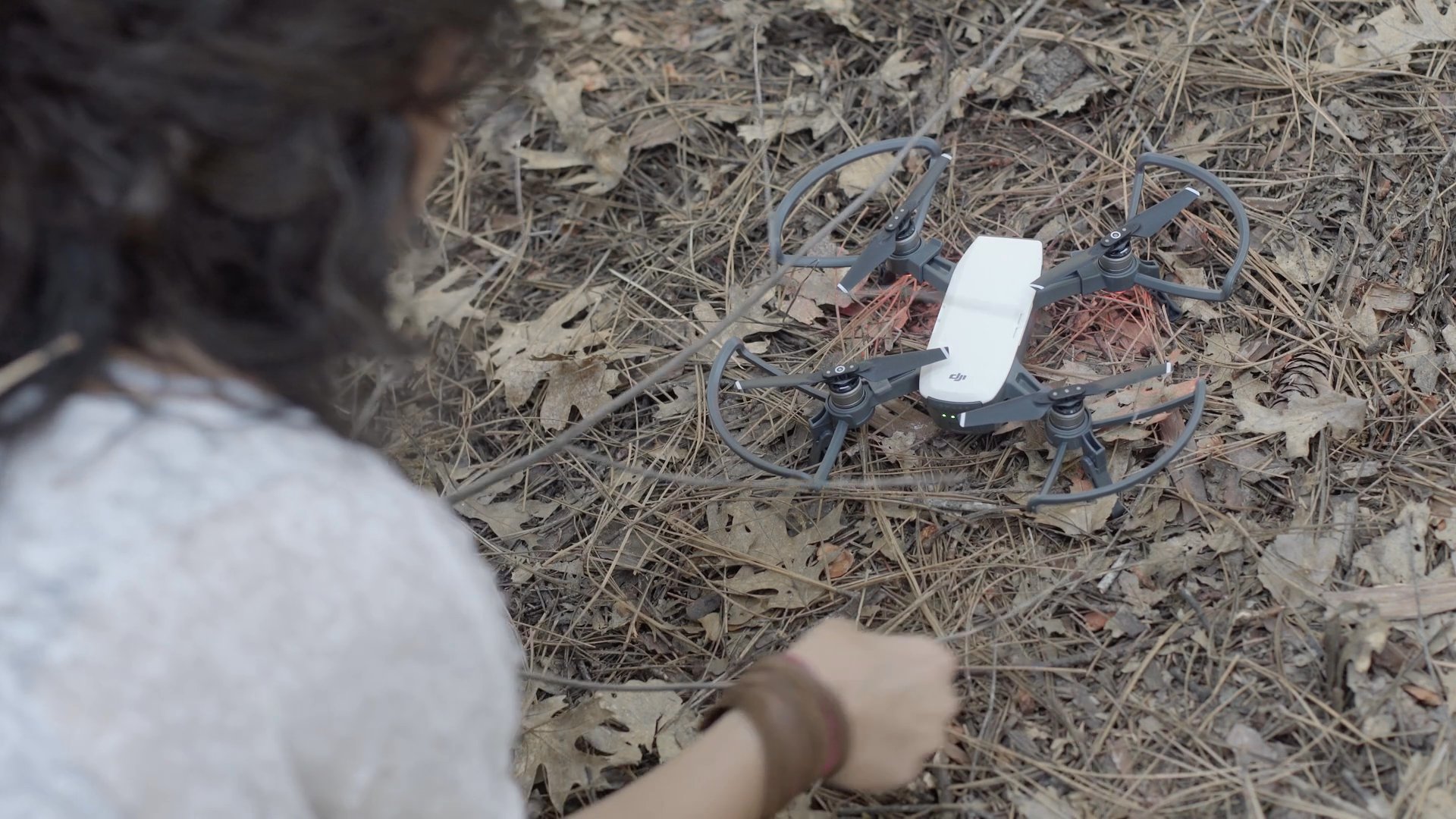 A woman prepares a small unmanned aerial vehicle on the forest floor before successfully launching it into the air. The drone hovers as the operator watches it rise from the woodland environment.