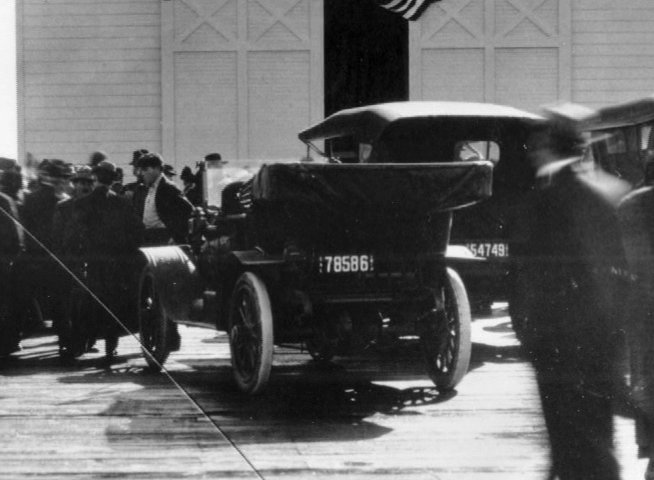 Crowds gather on a wooden pier alongside a large steamship as early 20th-century automobiles navigate the dock area. The scene documents maritime activity and port transit.