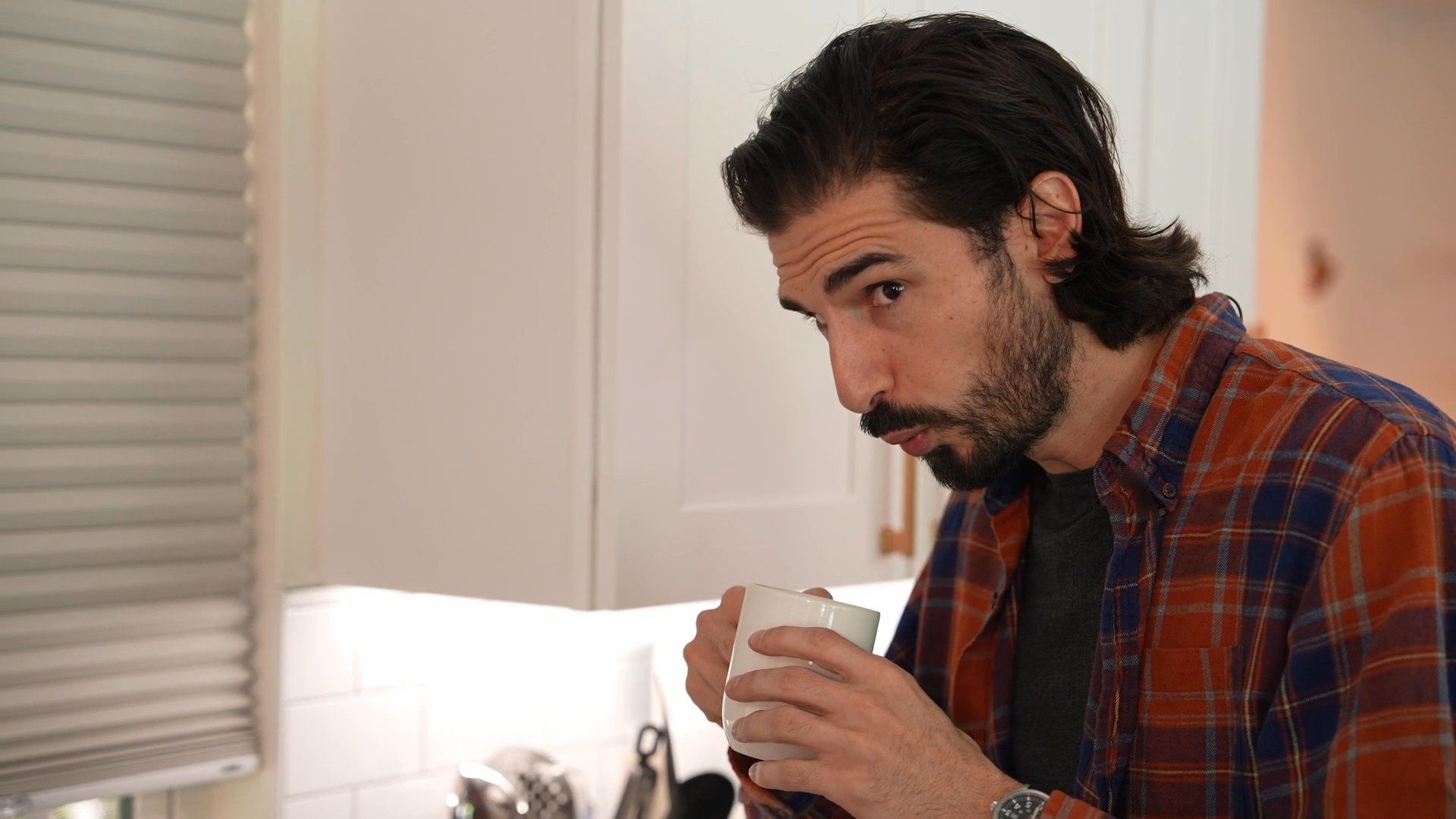 A woman prepares orange juice in a kitchen while engaging in conversation with a man who holds a mug. The two individuals interact casually within a domestic environment during their morning routine.