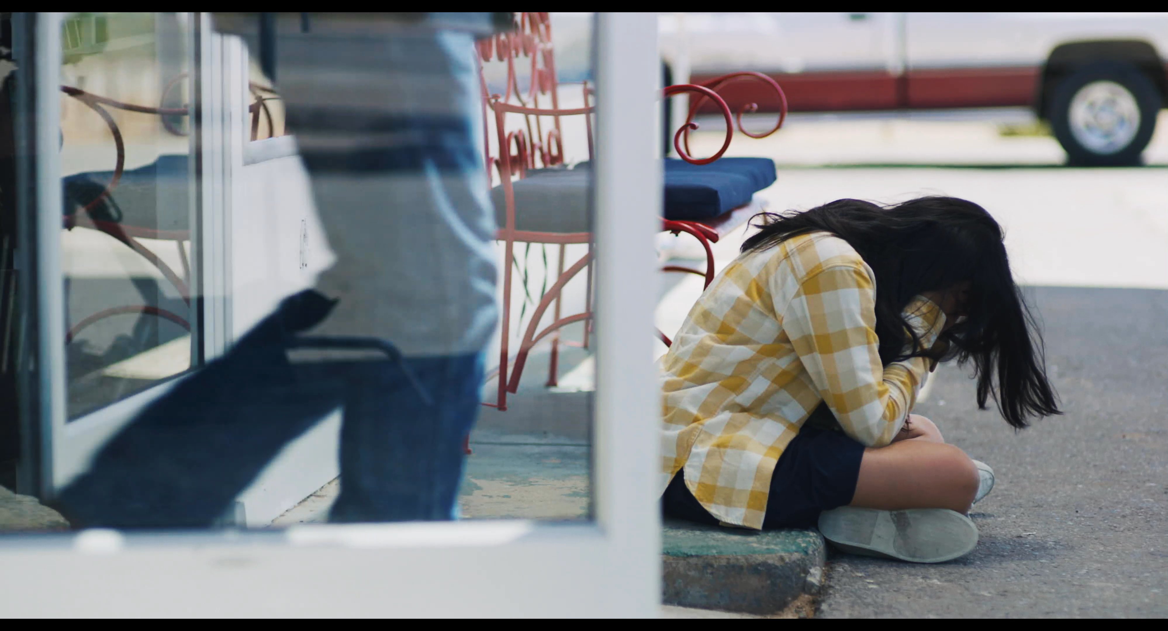 A woman in a yellow plaid shirt sits alone on a porch in distress, eventually receiving a drink from an individual who walks by.