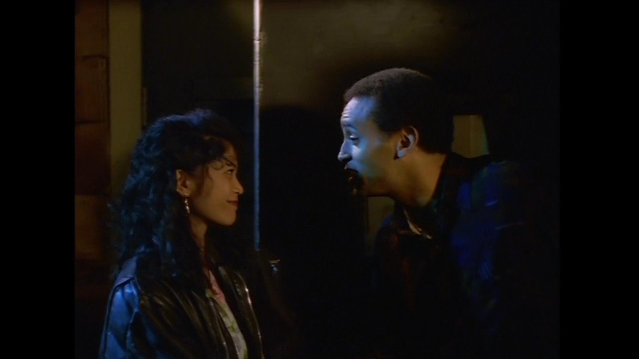 A man finishes a call at a public phone booth before engaging in a friendly conversation with a woman on a dim city street at night.