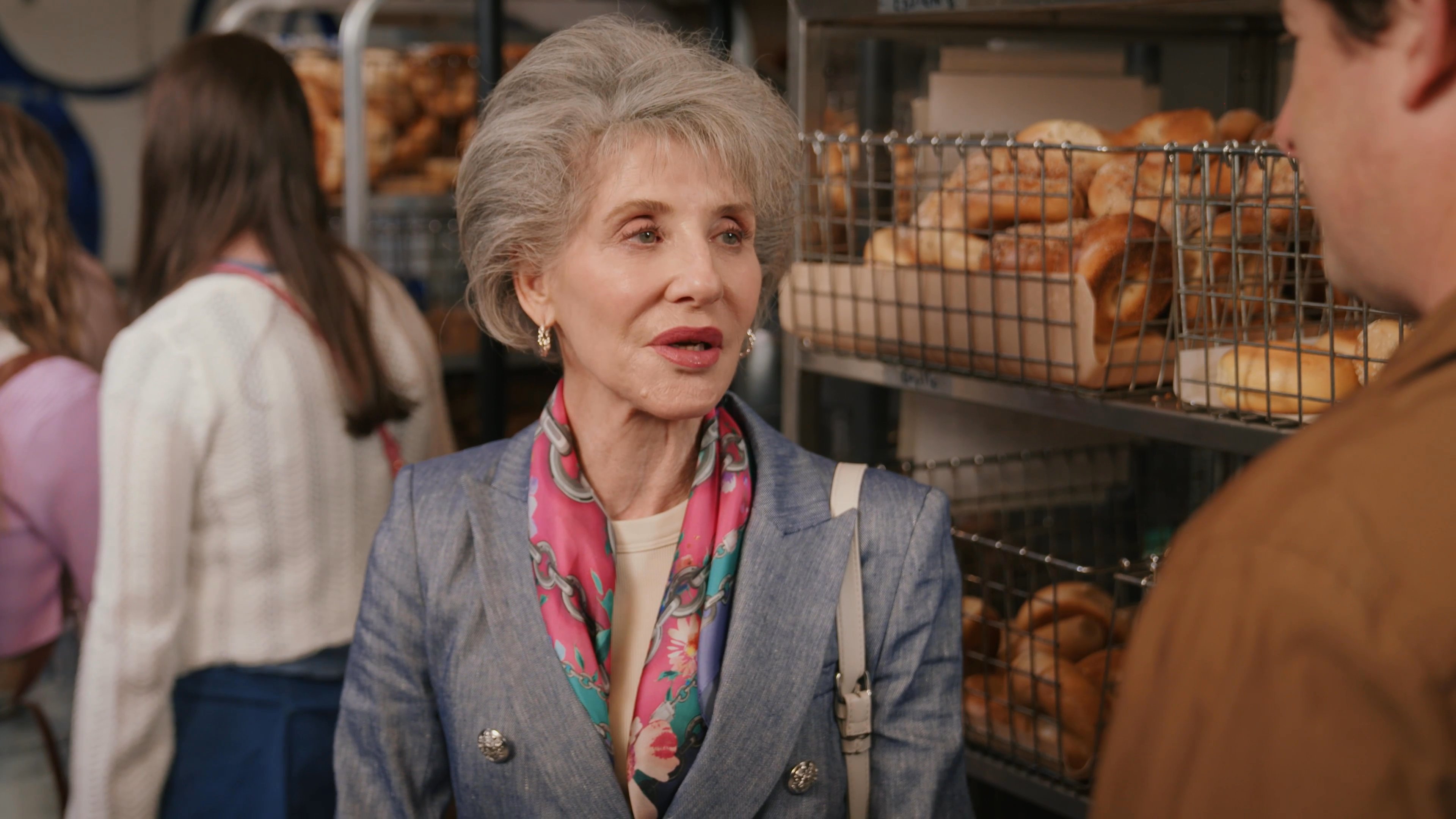 An elegant woman engages in a polite conversation with a man at a bakery counter filled with assorted bagels. The exchange reflects a professional or casual social interaction within a retail setting.