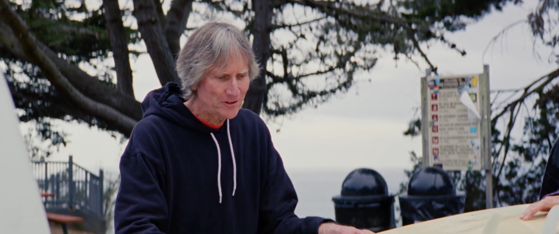 Three individuals gather at an oceanfront location to discuss and inspect surfboards. They exchange insights regarding surfing equipment while positioned near the water.