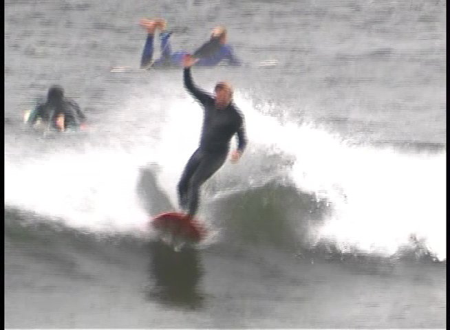 A surfer rides waves on a surfboard in the ocean while other surfers are visible in the background. The individual navigates various breaks with a balanced stance.