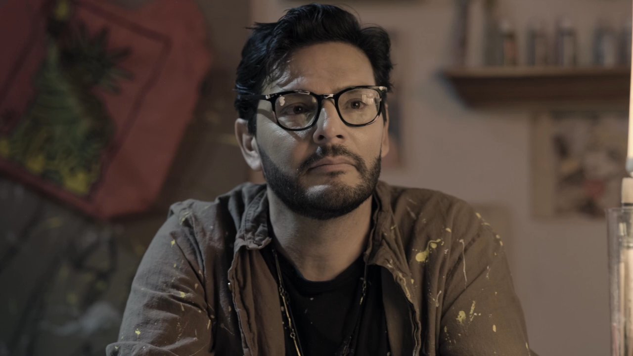 A man displaying a detective badge engages in a serious conversation with a woman within an artist studio. The interaction reflects a moment of inquiry or confrontation as the pair exchange words in the dimly lit space.