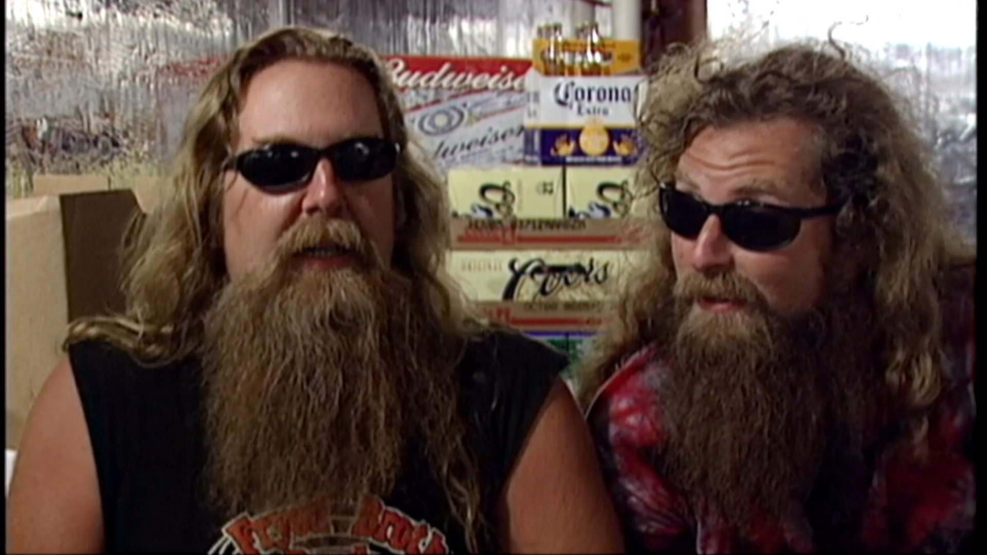 Two long-haired men with beards and sunglasses engage in an animated conversation during an indoor interview session.