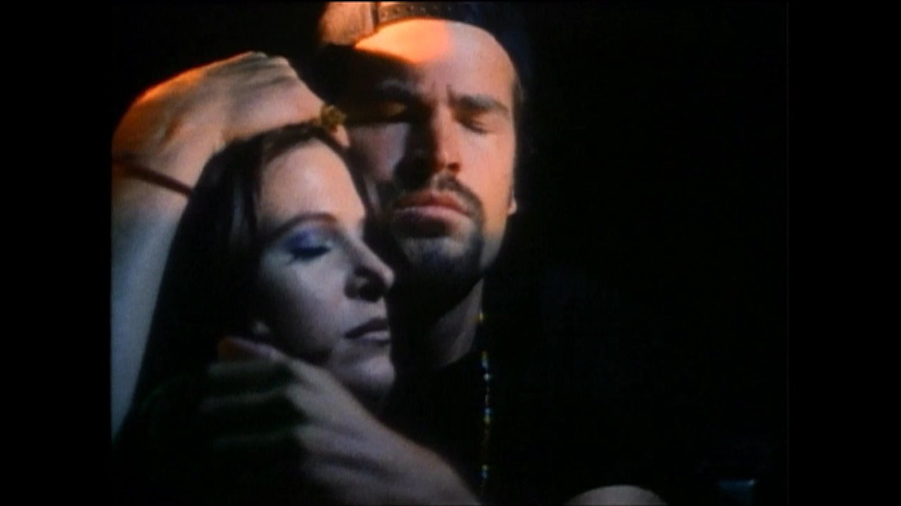 A man gently comforts and embraces a woman in a dimly lit, nocturnal setting, conveying a quiet moment of emotional connection.