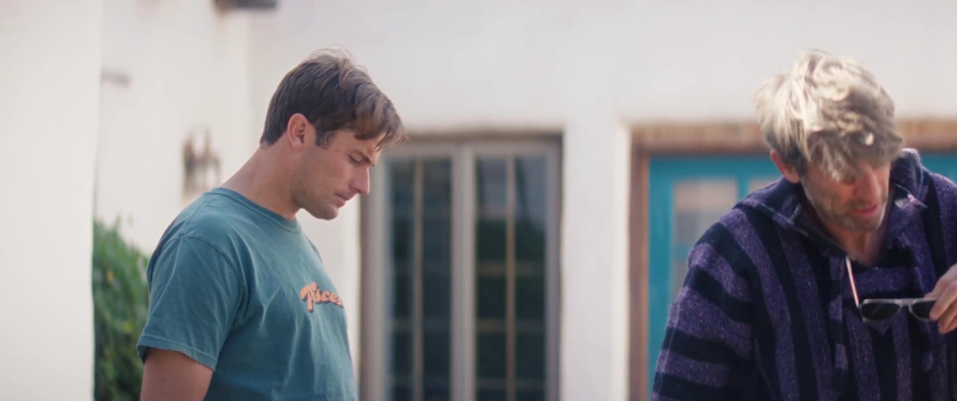 Two men engage in a conversation on an outdoor patio, shifting from a seated position to standing and walking together. The exchange reflects a casual social interaction in a residential setting.