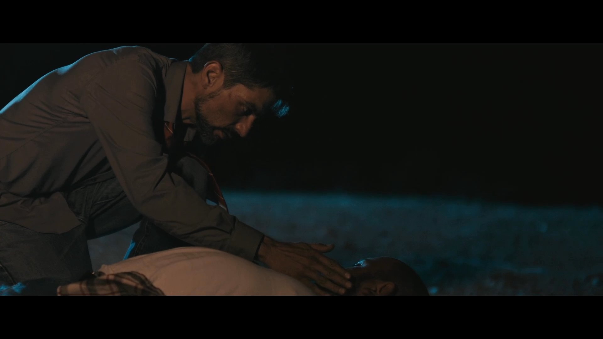 A man kneels on the ground at night to check on an unresponsive, bloodied individual lying on the dirt. He examines the person's condition with concern while wiping blood from his own hands.