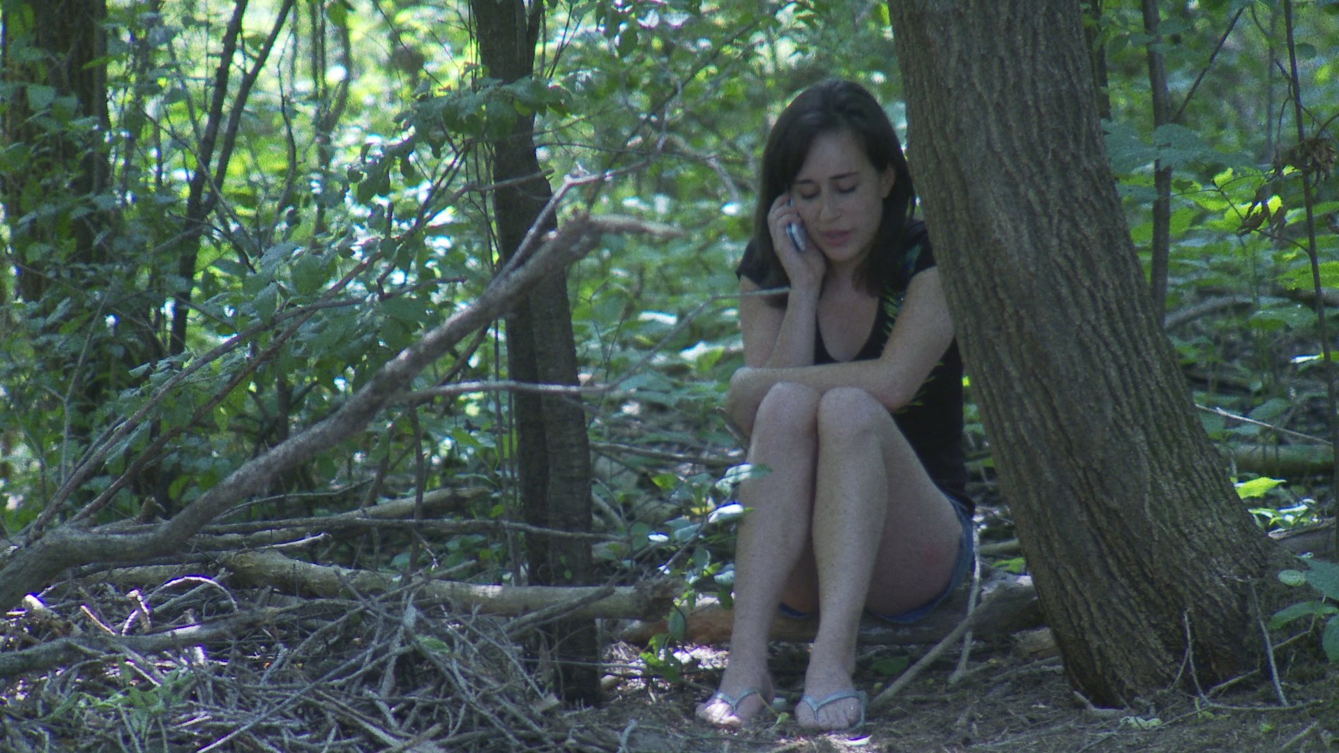 A woman walks a golden retriever through a wooded area while another woman sits near a tree, appearing upset during a phone conversation. The scene highlights the contrasting activities of pet care and personal crisis in a natural environment.