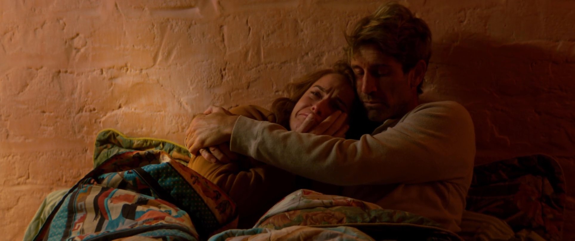 A man and woman share an intimate, supportive moment while cuddling under blankets in a dimly lit, warm-toned room. The woman appears emotional as the man holds her closely to provide comfort.
