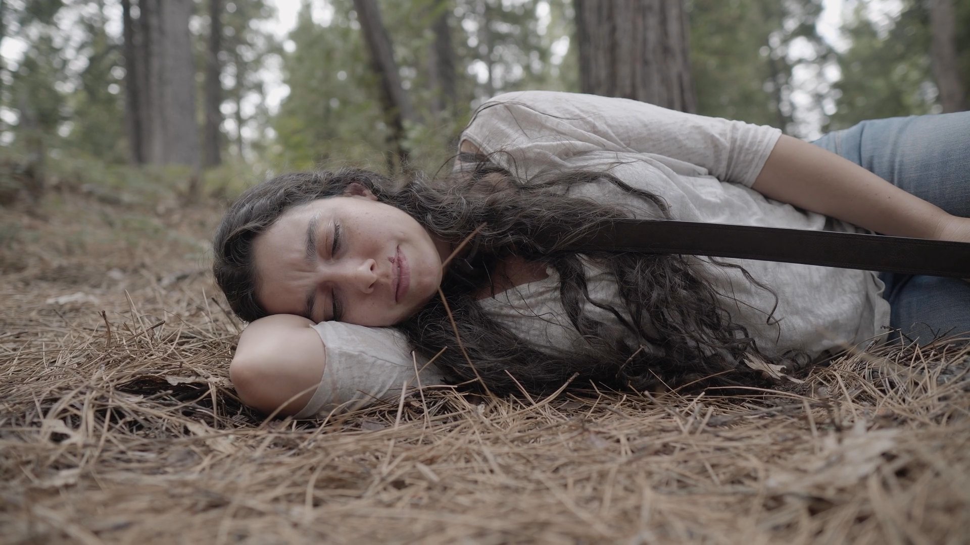 A woman with an injured, bandaged arm rests on the pine needle-covered forest floor, showing signs of physical distress and exhaustion.