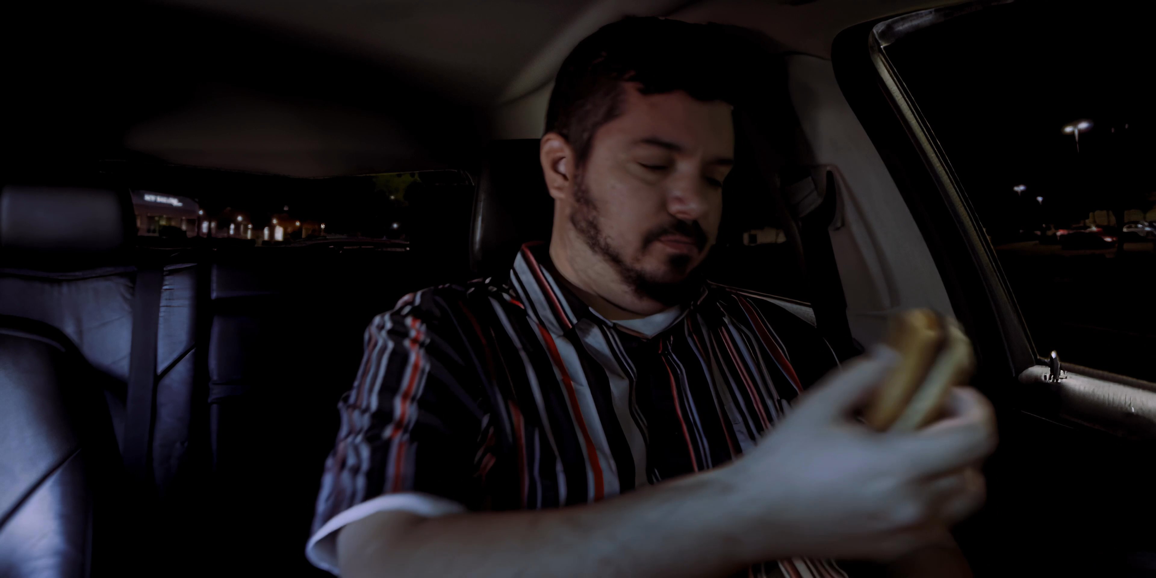 A man sits in the passenger seat of a vehicle at night and eats a hamburger. The setting is dimly lit, focusing on the individual during a solitary meal.