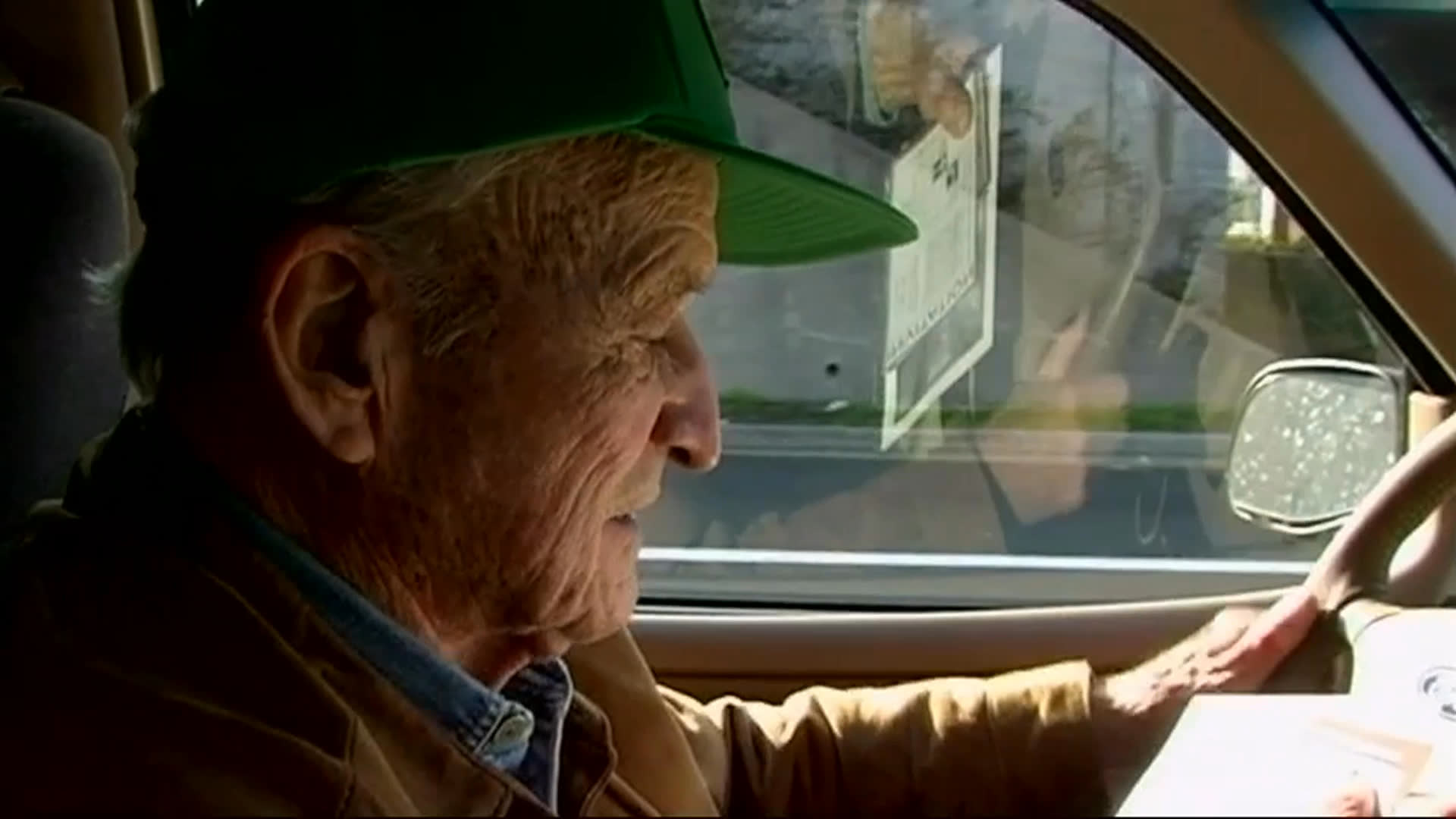 An older man in a green cap writes at a table, then drives through the city and arrives at an art gallery. He walks among the displayed works and chats with visitors in the bright exhibition space.