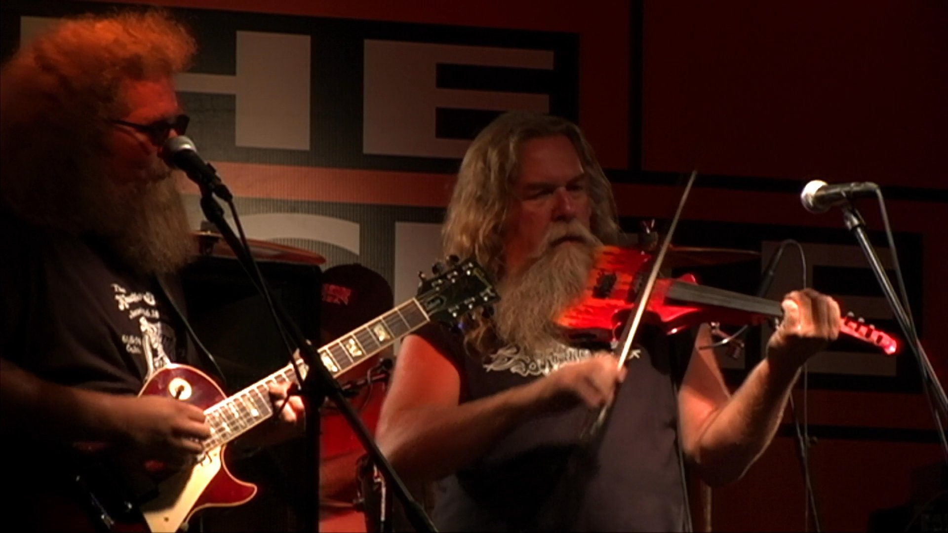 A band performs live music on a dimly lit stage in a bar, featuring a violinist and a guitarist entertaining a dancing crowd.